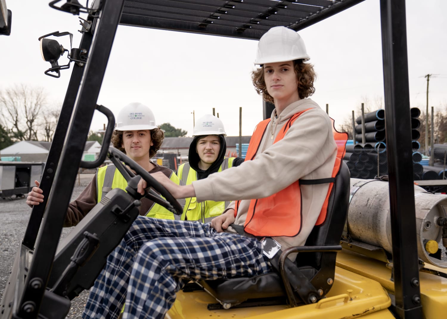 Student driving heavy machinery in hard hat, other students looking on